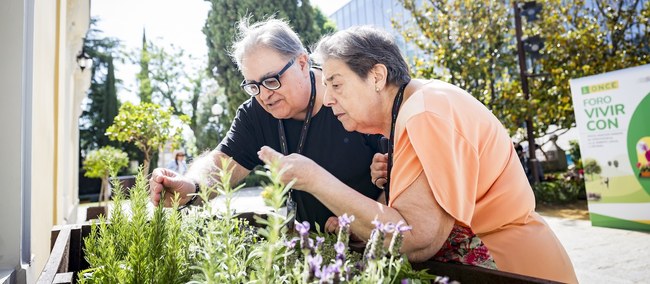 Dos personas mayores tocando plantas en un día soleado, al fondo letrero de Vivir con C