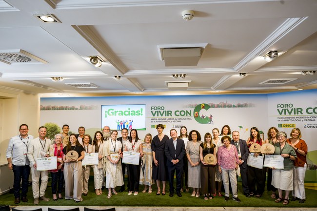 Gran foto de familia en el escenario con cartel detrás de Vivir con C 2026