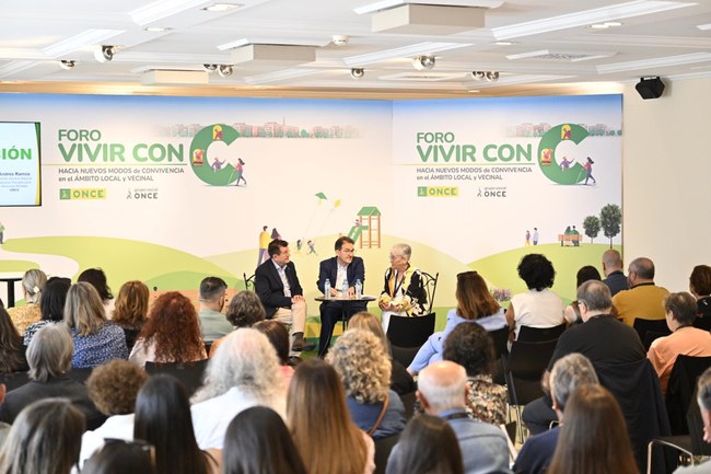 Foto del escenario con 3 participantes y público delante