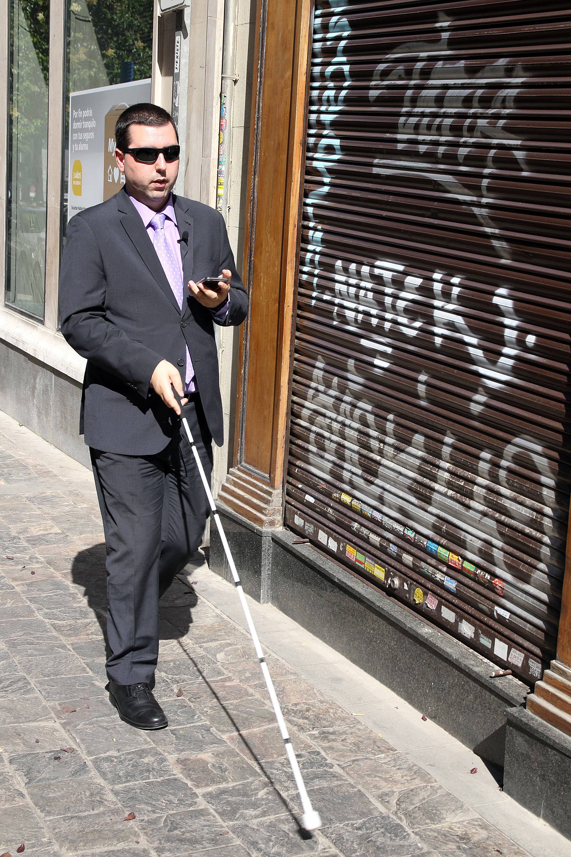 Una persona ciega camina con su bastón por la calle siguiendo las instrucciones de Moovit en su smartphone.