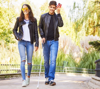 Una chica y un chico jóvenes caminan por el parque, él es ciego, lleva bastón y va oyendo un audio de Whatsapp
