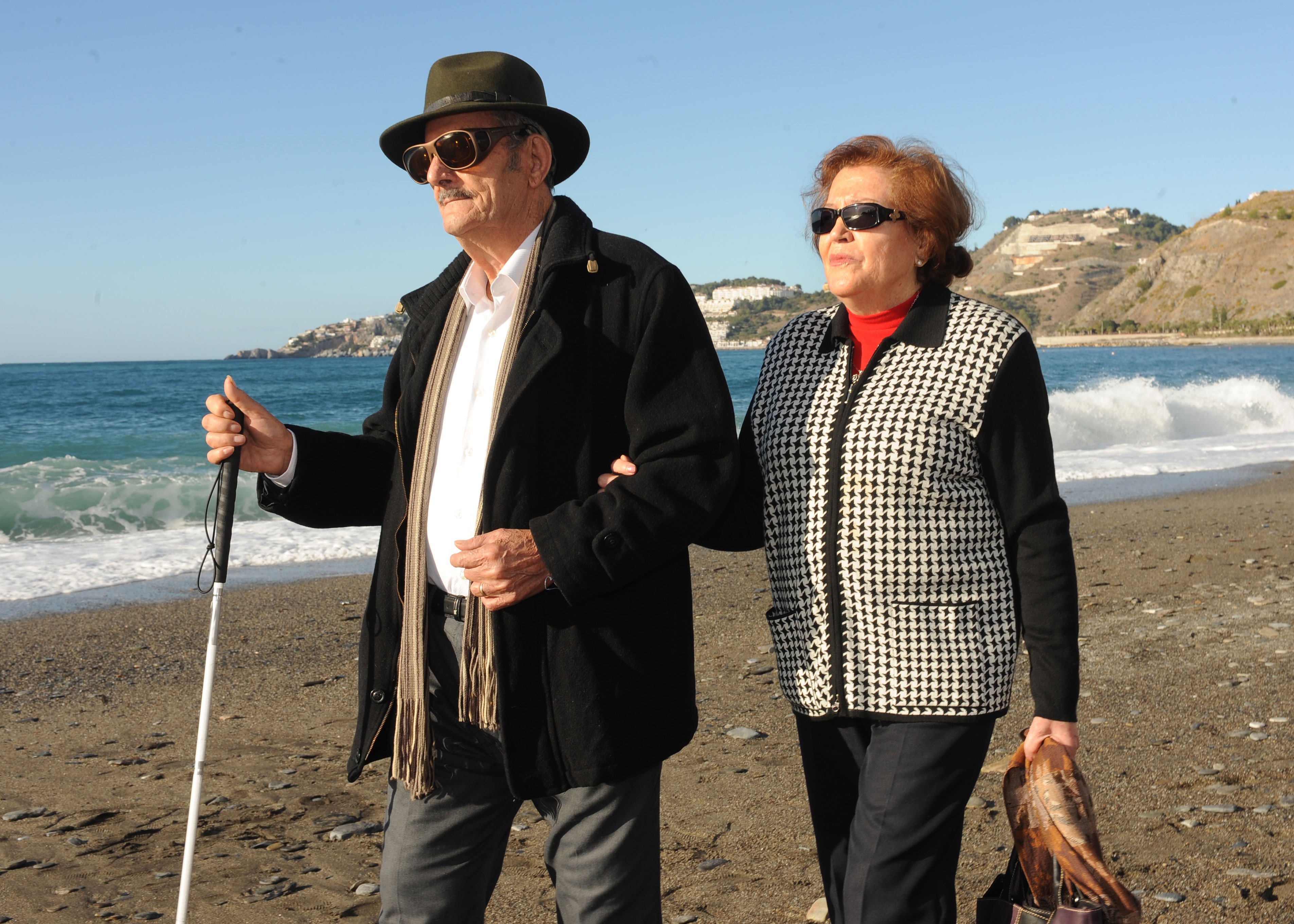 Un hombre y una mujer mayores caminando por la playa, él con bastón blanco