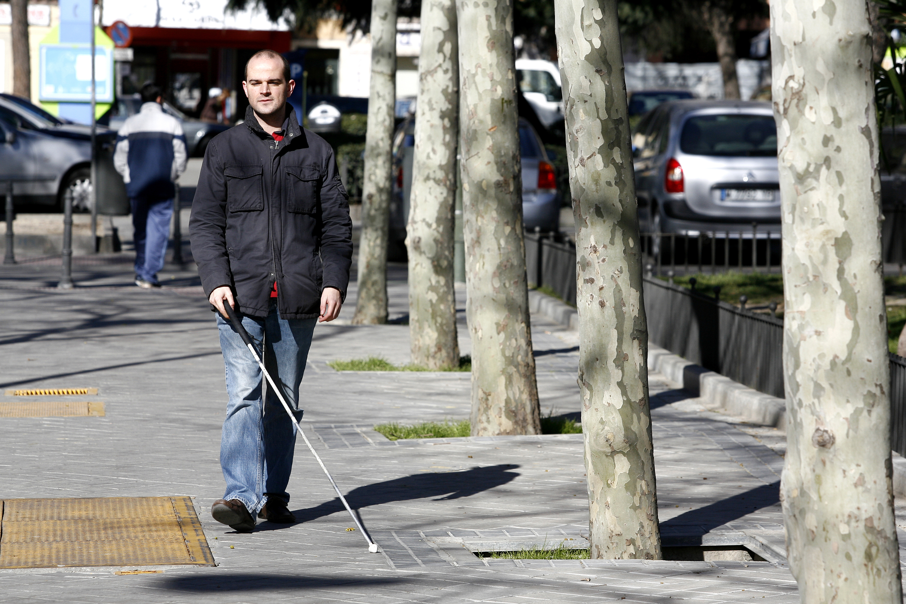 Un hombre ciego caminando por la acera con bastón blanco y árboles a la derecha